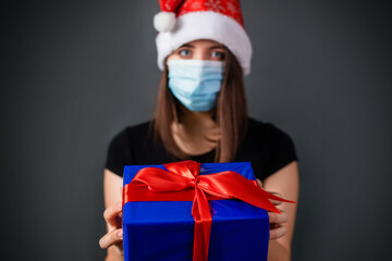 Fototapeta na wymiar Focus on a blue gift box with a red bow in the hands of th woman in a medical mask and a Christmas hat on a gray background in the dark.
