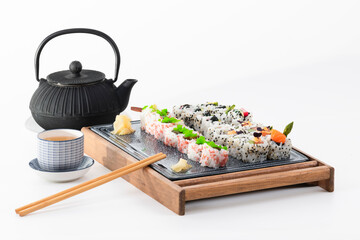 Tempting assorted uramaki pieces with chopsticks on a tray, with traditional cup and kettle on a light background.