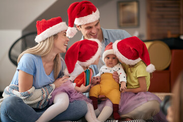 Fototapeta premium mionor sisters looking at baby brother, sitting in laps of young parents, wearing santa hats.