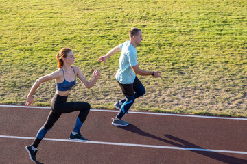 Obraz premium Young couple of fit sportsmen boy and girl running while doing exercise on red tracks of public stadium outdoors.