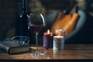 Glass of red wine, open book with candles in the background on wooden table.