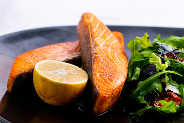 Delicious salmon with green salad and lemon on a black plate.