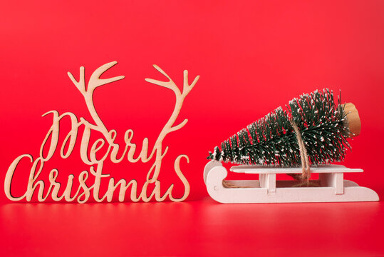 Christmas is coming concept. Close up photo of miniature christmas tree on little wooden sledge and merry chrismas wooden curved decoration isolated on red background