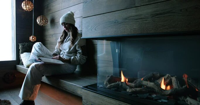 Woman reads a book after skiing, next to a fireplace. Attractive woman resting and looking at a book 4K