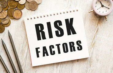 RISK FACTORS text on a sheet of notepad. Coins are scattered, pencils on a gray wooden background.