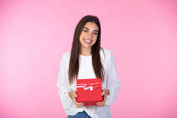 Smiling woman hold red gift box. Isolated pink background. Studio. Image of Happy woman in shirt holding gift box and looking at the camera over pink background