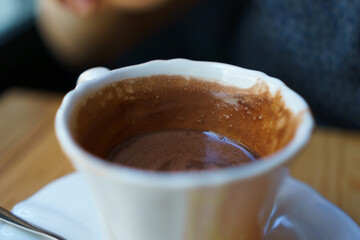 A white porcelain cup of hot chocolate or cocoa on a wooden table. On the saucer is a spoon and in the background you can see a blurred blue sweater and an arm.