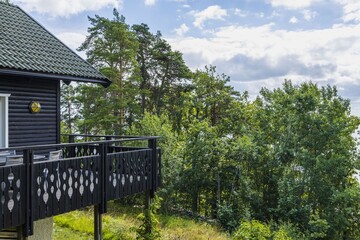 Fototapeta na wymiar Beautiful view of part facade of typical wooden Swedish house with wooden patio. Tops of green forest trees on blue sky and white clouds background on sunny summer day. 
