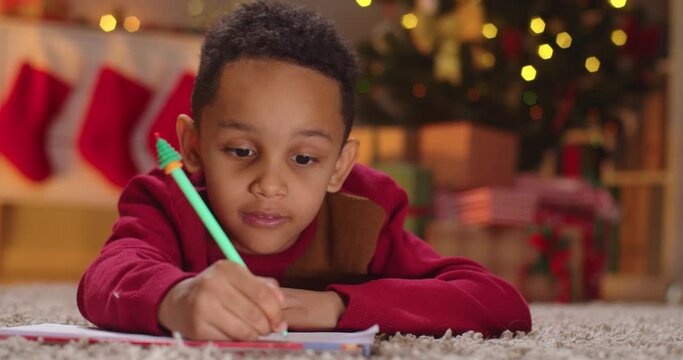 Close up of African American small kid boy on floor drawing and writing on paper in decorated room with blurred background. Little child writes letter to Santa. Xmas wishes concept