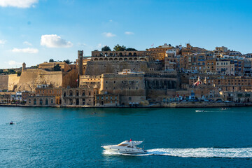 Fototapeta na wymiar A small yacht in the Grand Harbour in Malta passes the fortified capital city Valletta showing the Lascaris Battery, St. Peter and St. Paul Counterguard, Saluting Battery and Upper Barrakka Gardens.