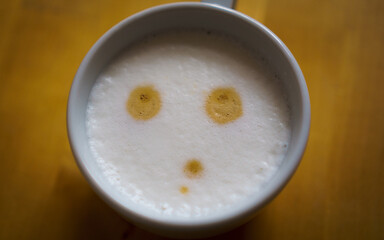 A cup of cappuccino coffee with milk foam showing a thoughtful face, on a blurred wooden table.