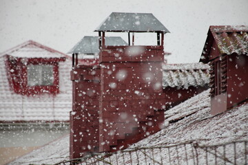 Winter snowfall in Europe, snowflakes on old city house with snow covered red tiled roof with stovepipes on gray sky background, European outdoor december landscape