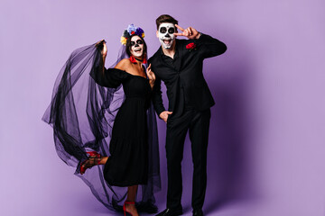 Full-length photo of funny guy and girl in masquerade masks laughing and posing in good mood. Lady in black veil with flowers in her hair touches boyfriend showing peace sign