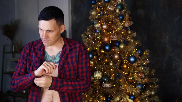 Handsome guy with unbuttoned shirt standing near Christmas tree in dark room. Young man buttons his shirt, preparing to celebration of Christmas.