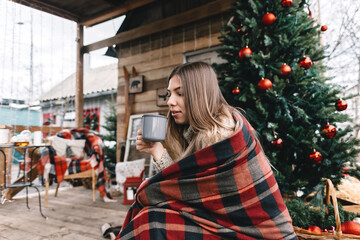 Young caucasian woman in a plaid is drinking a hot drink on the veranda with Christmas tree and decorations.