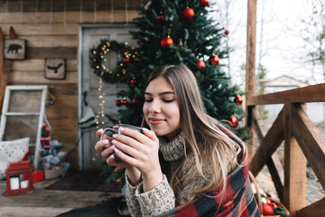 Young caucasian woman in a plaid is drinking a hot drink on the veranda with Christmas tree and decorations.