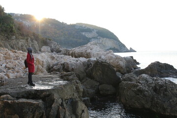 A woman wearing a headscarf in a red coat is watching the sunset while standing on the rocks by the sea