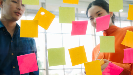 Business people work on project planning board in office and having conversation with coworker friend to analyze project development . They use sticky notes posted on glass wall to make it organized .