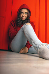 Young woman in hood sitting on floor. Curly brunette poses near red wall.