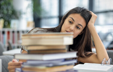 Beautiful frustrated female student looks at stack of books and holds her head while sitting at table in the university library. Studying and education concept
