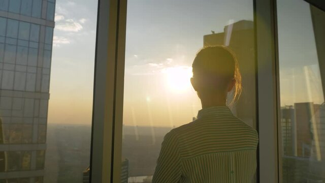 Back view of woman silhouette looking at cityscape through window of skyscraper. Summer time, sunset, sun lens flare. Success, business, opportunity, sightseeing, corporate, discover, future concept