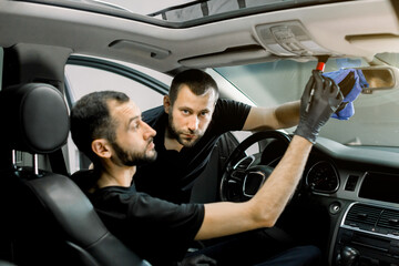 Two handsome male auto service workers cleaning modern car interior. Focus on the man, cleaning car mirror with microfiber cloth, while his colleague removes the dust from roof plastic panel