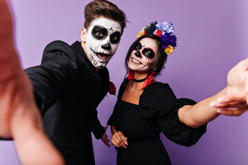 Funny young guy and his girlfriend make selfie with smiles on their faces. Portrait of naughty couple with Halloween makeup in purple studio