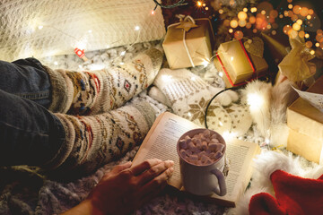 female legs in warm knitted socks on a plaid next to Christmas tree and decorations, a book and a cup of hot cocoa. atomsphere of holiday and comfort