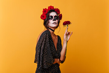 Charming female model in elegant dress enjoying scent of roses. Portrait of girl with Halloween makeup posing in orange room