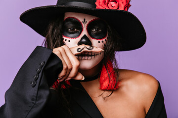 Mischievous brown-eyed girl with face art is having fun by putting painted mustaches to her mouth. Photo of stylish woman in black outfit with red details