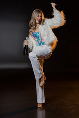 full length of blonde woman in faux fur jacket holding bottles with champagne on black