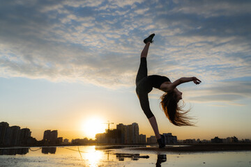 Female dancer showing her flexibility during sunset on cityscape background with reflection in the water. Concept of freedom and happiness 