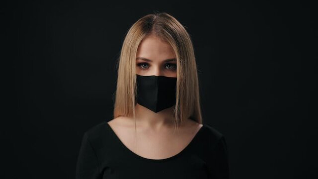 Attractive young woman with blond hair wearing black medical mask and sweater. Charming female posing in studio with black background.