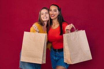 Two cheerful young brunette girlfriends in casual denim clothes posing while holding a shopping bag after shopping, isolated on red background.