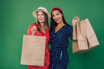 Cheerful two young women friends in dresses isolated on a green background in the studio. People lifestyle concept. Keep a bag after shopping.