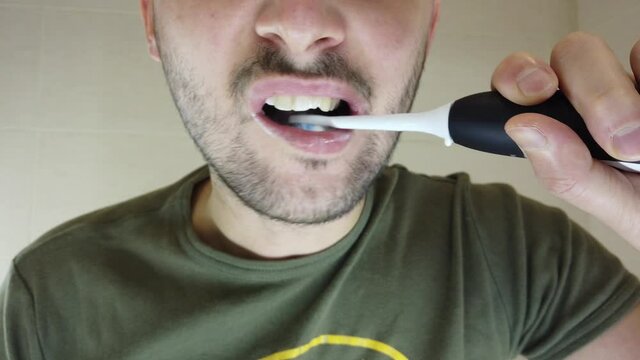 A bearded man brushes his teeth in the mirror with his mouth open. Toothpaste on an electric toothbrush