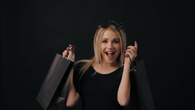 Happy young woman with blond hair looks satisfied with successful shopping. Smiling female holding paper bags over black background.