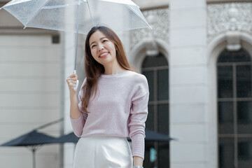 Fototapeta na wymiar Young Asian woman smile and holding a transparent umbrella under the rain in the city