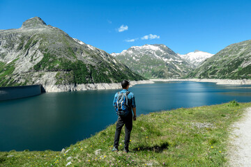 Fototapeta na wymiar Man standing at the side of a lake at Kölnbreinsperre dam in Austria. The lake has navy blue color. High Alps around. There is a glacier in the back. The meadows blossoming with wild flowers. Calmness