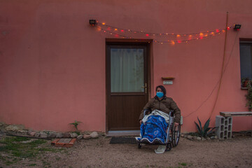 elderly in wheelchair with health mask front of house with christmas lights