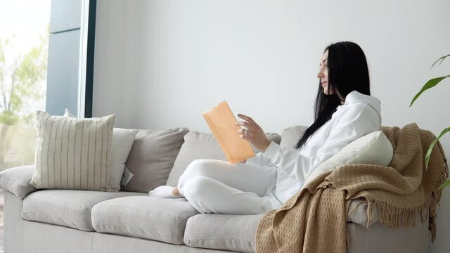 Young woman reading a book while sitting on a sofa in a living room, reading books. Stay home concept