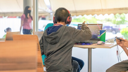 Back view of Asian boy wearing protective face mask and headphones while working with a friend on laptop during corona virus (Covid-19) and flu outbreak in food court at the mall.New normal lifestyle.