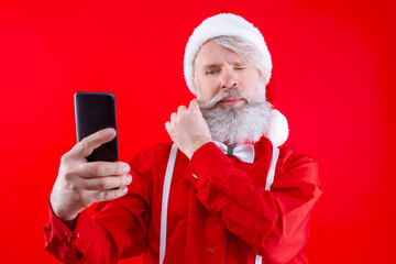 Happy modern Santa Claus, wearing stylish red hat, shirt and suspenders. Positive, old man with long white beard doing selfie on smartphone and smiling. Men holding his mustache and shopping, online.