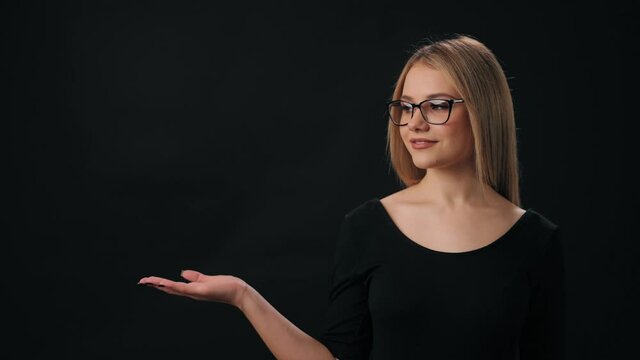 Front view of charming woman in eyeglasses holding empty palm over black background. Space for text and advertisement. Black friday concept.