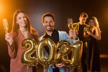 multicultural couple holding glasses of champagne and balloons with 2021 numbers near friends on blurred background on black