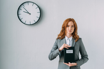Redhead female lawyer holding book with intellectual property lettering, while standing near wall...