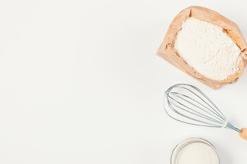 Top view of bag of flour next to whisk and sugar on white kitchen table