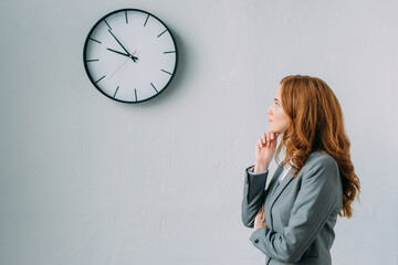 Side view of thoughtful businesswoman looking at wall clock on grey, stock image
