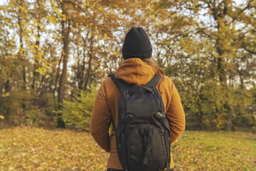 Woman in a yellow jacket with her hends in her pockets and a backpack on her back walking trough the woods in the fall
