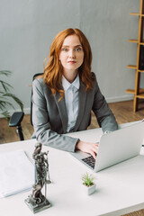 Redhead female lawyer looking at camera, while typing on laptop at workplace with blurred themis figurine 